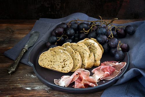frisches Vollkornbrot mit blauen Trauben und Prosciuttoschinken auf einem dunklen rustikalen Holztis