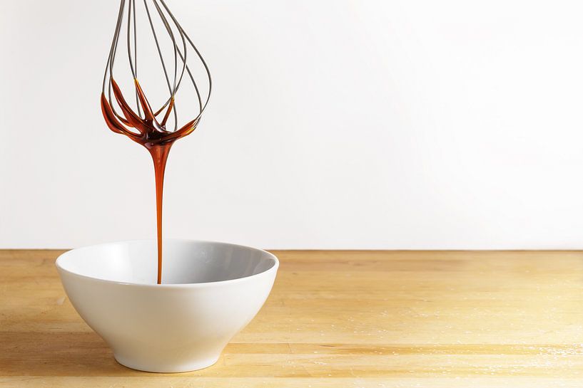 Brown sugar syrup flows from a wire whisk into a white bowl, wooden table and bright background with by Maren Winter