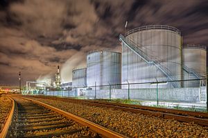 Öl-Raffinerie in der Nacht mit Schienen und Silos gegen einen bewölkten Himmel von Tony Vingerhoets