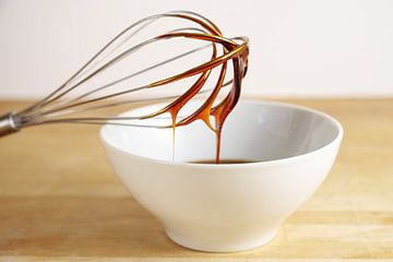 Brown molasses flows from a wire whisk into a white bowl, baking at home concept, wooden table and b by Maren Winter