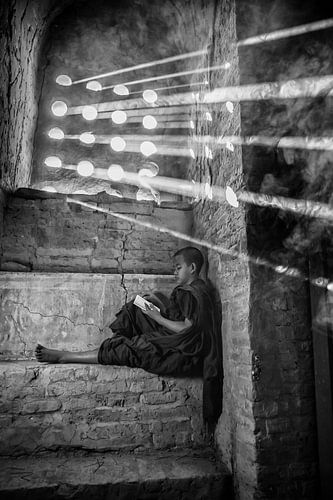 Young monk reads prayer book sitting on a windowsill of a monastery in Baghan with strong sunshine
