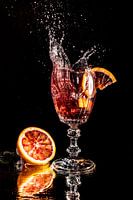 Blood orange piece falls into a glass with water mirrored on black background