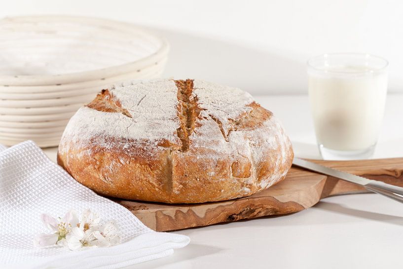 Homemade sourdough bread on a wooden board, with proofing basket, knife, gallon milk against a light by Ans van Heck