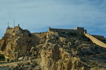 Het Castillo de Santa Bárbara in Alicante op een rotsachtige berg onder een strakblauwe hemel. van LuCreator