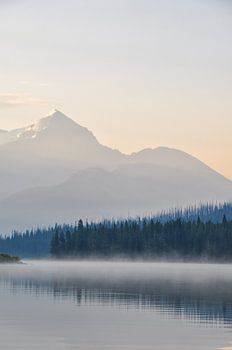 Ein ruhiger See in Kanada, umgeben von majestätischen Bergen und üppig grünen Bäumen, unter einem blauen Himmel bei Sonnenaufgang, mit Tau auf der Wasseroberfläche. Das ruhige Wasser spiegelt die Landschaft wider und schafft ein Gefühl von Symmetrie und Gleichgewicht. Die Landschaft strahlt Ruhe und Gelassenheit aus. Dies ist ein Foto des Maligne-Sees in Kanada. Der Sonnenaufgang verleiht ihm schöne gesättigte Farbtöne.