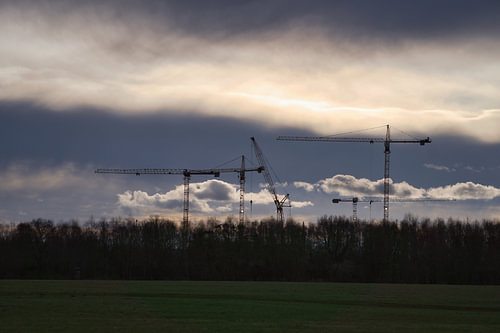 Baukräne ragen während eines dramatischen Sonnenuntergangs über einer bewaldeten Landschaft auf.