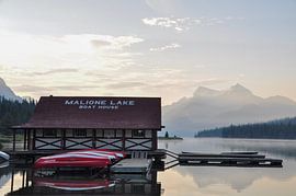 A serene lake in Canada, surrounded by majestic mountains and lush green trees, under a blue sky during sunrise, with dew on the surface of the water and a boathouse in the foreground. The calm water reflects the landscape, creating a sense of symmetry and balance. The landscape exudes peace and serenity. by UMA Digital NL