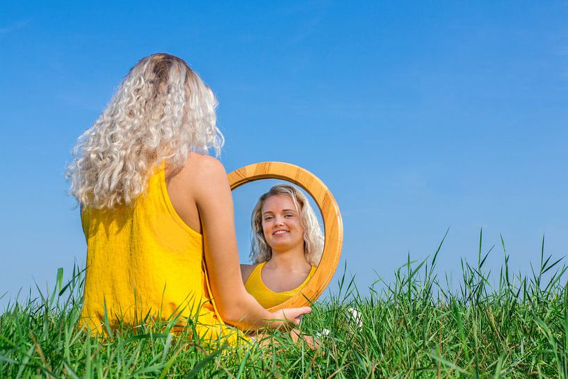 Jonge vrouw zit met spiegel in gras met blauwe lucht van Ben Schonewille