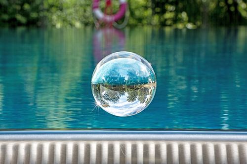 Glazen bol zweeft boven een zwembad met een spiegelende bewolkte lucht en bomen