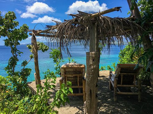 Blick auf eine sonnige Lounge mit Bambusstühlen, Palmendach, im Hintergrund das Meer im asiatischen Raum