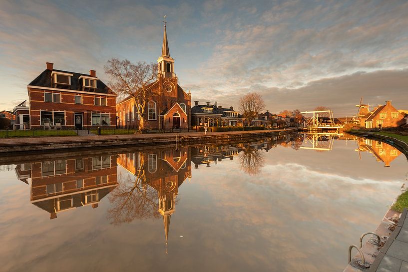 n the warm light of the setting sun. The soft hues of orange, gold and pink fill the sky and give the mill a fairytale feel. The light subtly plays on the details of the mill and the surrounding nature, while the reflections in the water enhance the serene atmosphere. The composition and the warm play of colours are reminiscent of a classical painting, which focuses on tranquillity and romance. This photo captures the essence of the Frisian landscape and invites you to dream away at the beauty of Birdaard's church and windmill reflected in the water at sunset by KB Design & Photography (Karen Brouwer)