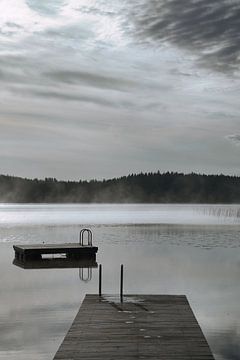 A wooden jetty on the lake in the morning mist, flooded with sunlight and a calm reflection. by Martin Köbsch