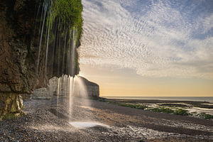 Ein natürlicher Wasserfall entlang der Kalksteinfelsen der Normandie bei Sonnenuntergang und Ebbe im goldenen Abendlicht, mit einer beeindruckenden Küstenlinie und einem spektakulären Wolkenhimmel