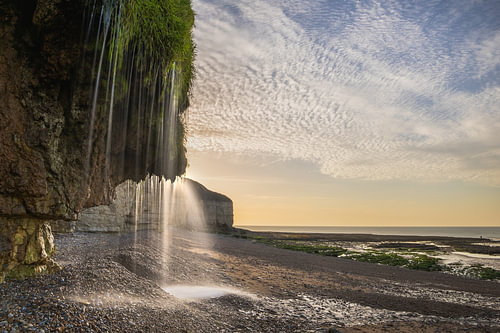 Natuurlijke waterval langs de kalkstenen kliffen van Normandië tijdens zonsondergang bij eb met gouden avondlicht, indrukwekkende kustlijn en spectaculaire wolkenlucht