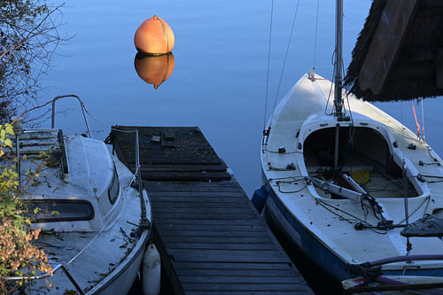 Zwei schmutzige Boote an einer Anlegestelle und eine orangefarbene Boje im blauen, ruhigen Wasser ei