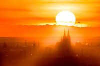 De blik dwaalt af naar de verte en daar, te midden van de gouden sluier van de ondergaande zon, rijst de Dom van Keulen op. Dit perspectief vangt zowel de uitgestrektheid van het landschap als de imposante aanwezigheid van de kathedraal.