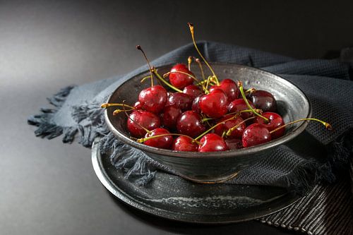 Fresh red cherries in a rustic ceramic bowl with napkin and plate on a dark gray background, healthy