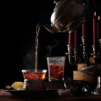 pouring hot steaming tea from a vintage teapot into  glass cups on a rustic table setting with candl