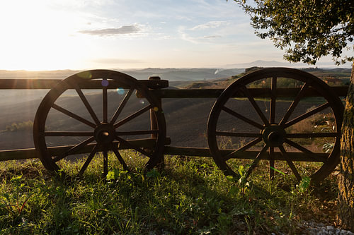 Old wooden wheels are leaning against a fence overlooking the Tuscan countryside
