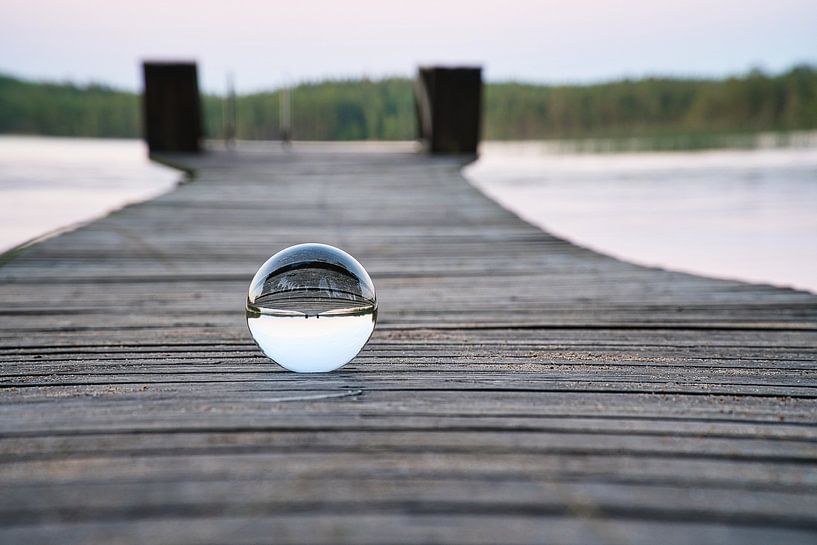 Glass ball on a footbridge reaching into a lake by Martin Köbsch