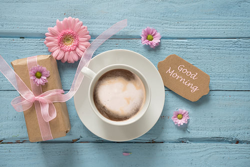 Kaffeetasse, rosa Blumen und ein Geschenk auf pastellblauem Holzgrund mit Kopierfeld, Text Guten Mor