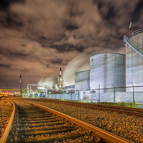 Oil refinery at night with rails and silos against a cloudy sky