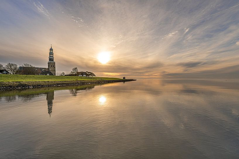 Sunset over the IJsselmeer and the tower of Hindeloopen mirrored in the still water. by Harrie Muis
