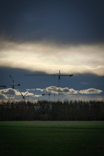 Baukräne ragen während eines dramatischen Sonnenuntergangs über einer bewaldeten Landschaft auf.