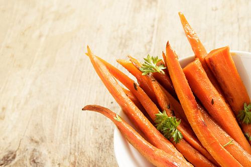 glazed carrot sticks  with parsley garnish in a bowl on a rustic wooden table, corner background wit