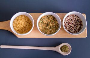 Houten plank met quinoa, rijst en couscous in witte schaaltjes - houten lepel met kruiden von Malu de Jong
