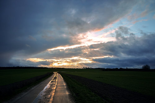 Nass reflektierende Landstraße aus Betonplatten führt durch die dunklen Felder unter einem dramatisc
