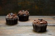 three chocolate cupcakes with cocoa butter cream on dark wood, one is in focus, copy space, narrow d by Maren Winter thumbnail