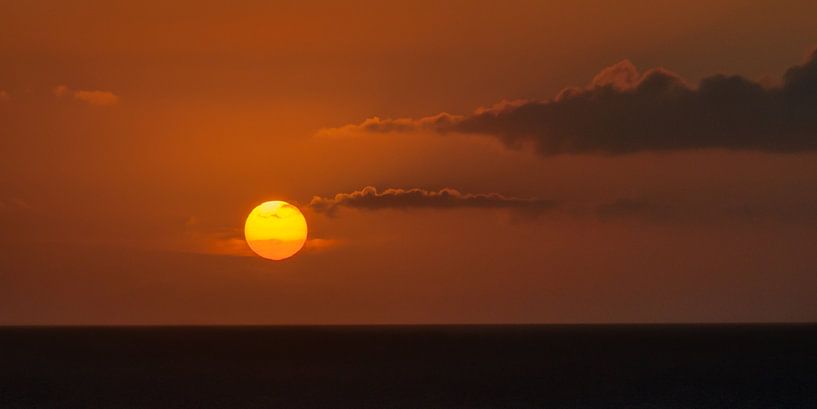 De zon tijdens zonsondergang en enkele wolkjes boven zee van Harrie Muis