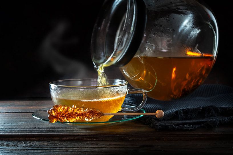 Du thé chaud et fumant est versé d'une théière dans une tasse en verre, servi avec un bâton de sucre par Maren Winter