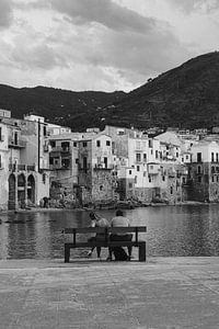 Paar an der Uferpromenade mit Blick auf die Stadt Cefalu, Sizilien Italien in schwarz und weiß von Manon Visser