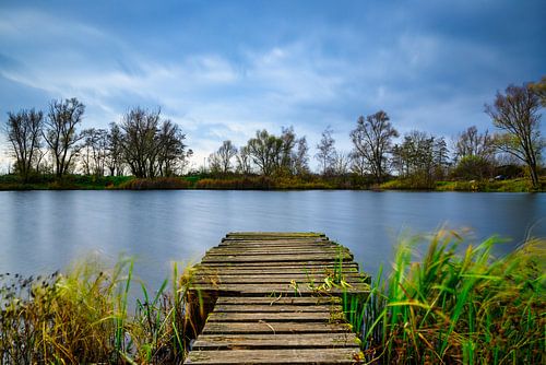 Het prachtige blauwe meer met bomen en de bewolkte lucht op de achtergrond en een houten pad dat er naartoe leidt