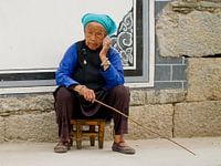 Portret van een chinese vrouw op een krukje met een stok in de hand voor een stenen muur in Dali