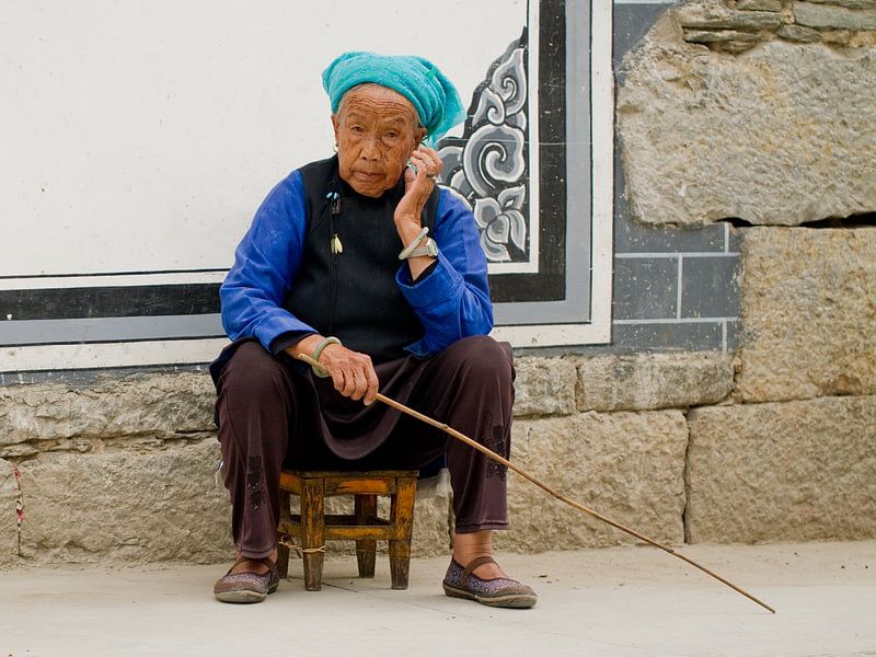 Portrait of a Chinese woman on a stool holding a stick in front of a stone wall in Dali by Cindy Mulder