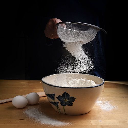 meel dat in een boog van een zeef in een kom op een houten keukenblad valt bij het bakken, donkere a