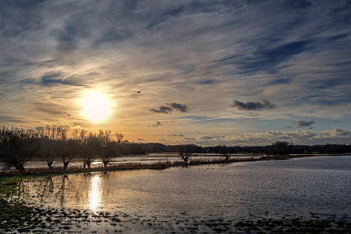 Untergehende Sonne mit Spiegelung im Wasser und bewölktem Himmel über einer dunklen Auenlandschaft a