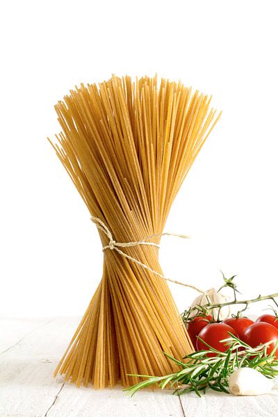 bosje volkoren spaghetti dat zich op wit hout bevindt met wat tomaten en kruiden die tegen een witte van Maren Winter