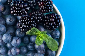 Blueberries and blackberries on a plate against a bright blue background. by Ans van Heck