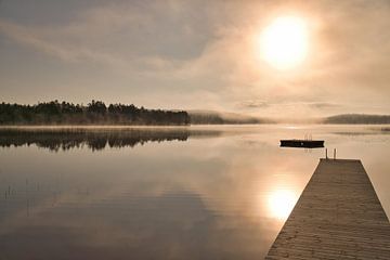 A wooden jetty on the lake in the morning mist, flooded with sunlight and a calm reflection. by Martin Köbsch