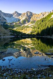 Ein idyllischer Bergsee liegt still eingebettet zwischen den schroffen Felswänden des Karwendelgebirges. Kristallklares Wasser, umgeben von alpinen Wiesen, Lärchen und der wilden Bergkulisse.