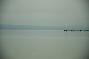 A long lonely footbridge reaches into the foggy Ammersee in Bavaria. Mountain ranges on the horizon