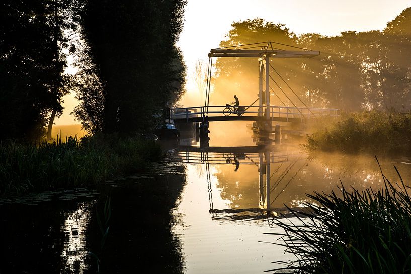 Lone newspaper delivery man cycles across a bridge in IJlst Friesland. One2expose Wout Kok Photograp by Wout Kok