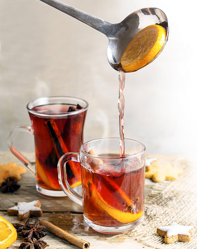 Gießen von Glühwein in Glasbecher, Weihnachtsplätzchen und Gewürze wie Orangenscheiben, Nelken, Ster