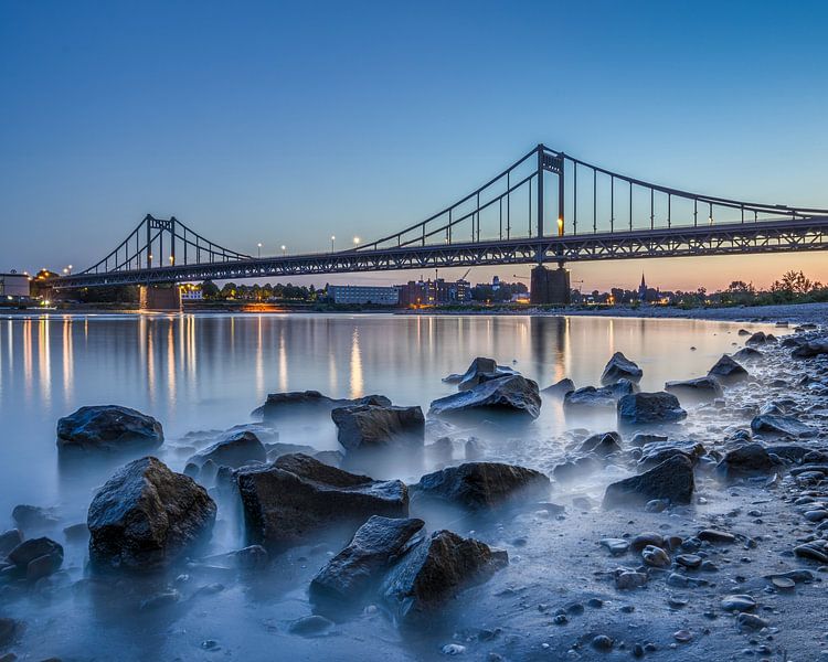 Die Krefeld-Uerdinger Brücke in der blauen Dämmerung, fotografiert vom Ufer mit seinem typischen Rheinkiesel. Dieses Panorama fängt die besondere Atmosphäre der blauen Stunde auf unvergleichliche Weise ein. von LICHTERKISTE