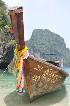 Un bateau traditionnel thaïlandais en bois flotte sur les eaux calmes d'une plage de sable dans une baie tropicale thaïlandaise, avec des montagnes verdoyantes en arrière-plan.