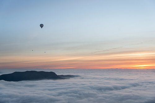 Three hot air balloons flying over a sea of ​​clouds during sunrise.